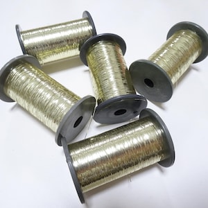 May include: Five spools of metallic gold thread are arranged on a white surface. Each spool has a dark grey core and edges, with the gold thread wound tightly around the core. The spools are angled in different directions.