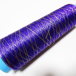 May include: A spool of vibrant purple thread with gold metallic accents. The thread is wound around a light blue plastic core. The gold threads create a crisscross pattern across the purple thread.