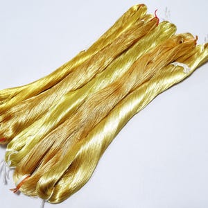May include: A collection of golden-yellow silk threads, bundled together. The threads have a shiny, smooth texture and are tied with small red and white strings. The image is set against a plain white background.