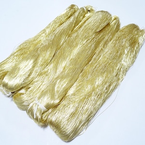 May include: Several bundles of golden-yellow silk thread are arranged on a white surface. The silk has a soft, shimmering appearance, with individual strands visible. The bundles are tied together with white thread.