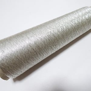 Könnte beinhalten: Eine Spule silberfarbenes Metallic-Garn. Der Faden ist um einen Pappkern gewickelt und bildet eine zylindrische Form. Der metallische Glanz verleiht dem Faden ein schimmerndes Aussehen, das Licht reflektiert. Die Spule ist etwa 10 cm lang.