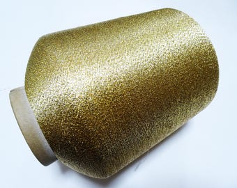 May include: A spool of gold metallic thread. The thread is tightly wound around a cardboard core, creating a cylindrical shape. The thread has a shimmering, textured appearance, reflecting light.