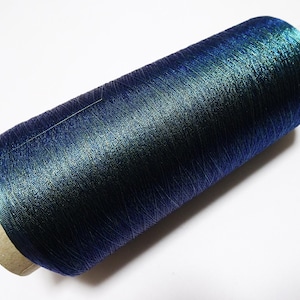 Op de afbeelding: Een klos garen met een levendige, kleurveranderende afwerking die varieert tussen diep marineblauw en iriserend smaragdgroen. Het garen is strak gewikkeld rond een cilindrische kartonnen kern met zichtbare uiteinden.