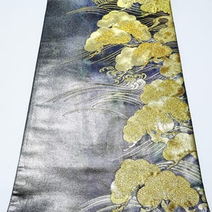 Può includere: Un tessuto rettangolare con sfondo grigio scuro e blu, con motivi ricamati dorati di nuvole e onde. Il tessuto ha una lucentezza metallica e il design è concentrato su un lato.