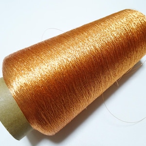 Puede incluir: Una bobina de hilo metálico naranja con una etiqueta de papel marrón.