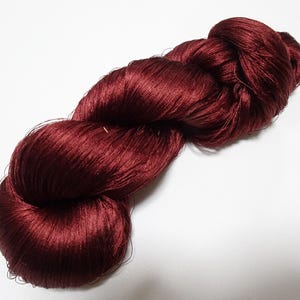 May include: A skein of deep burgundy yarn, tightly wound and glistening. The yarn appears to be silk or a similar luxurious fiber, with a smooth, reflective surface. The color is a rich, saturated red, perfect for knitting or crocheting projects.