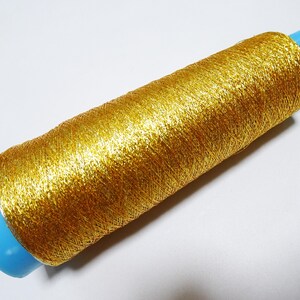 May include: A spool of gold metallic thread. The thread is tightly wound around a light blue plastic core. The metallic thread has a shiny, textured appearance, reflecting light. Ideal for crafting and embellishments.