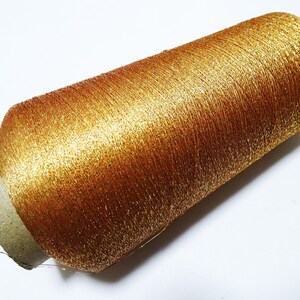 May include: A spool of metallic gold thread. The thread is tightly wound around a cardboard core, creating a cylindrical shape. The thread has a shiny, textured appearance, reflecting light and giving it a luxurious look. Ideal for embroidery and crafting.