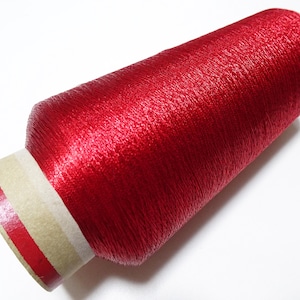 May include: A spool of red metallic thread. The thread is wound around a cardboard spool with a white label and a red stripe.