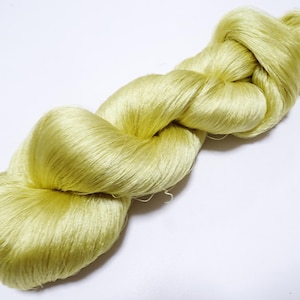 Könnte beinhalten: Ein Strang hellgelber Seidengarn, fest verdreht und geknotet. Das Garn hat einen weichen, glänzenden Glanz, was auf ein hochwertiges Material hindeutet. Die Farbe ist ein helles, gedämpftes Gelb, und das Garn scheint für verschiedene Textilprojekte bereit zu sein.