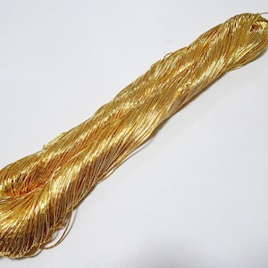 Może przedstawiać: Pęczek metalicznej złotej nici, ciasno zwiniętej i ułożonej po przekątnej na białym tle. Nić składa się z wielu cienkich pasm, odbijających światło i tworzących efekt połysku. Materiał ten nadaje się do różnych projektów rzemieślniczych.