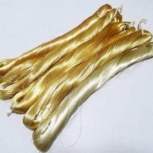 Puede incluir: Un conjunto de seis madejas alargadas de hilo de bordar de color amarillo dorado brillante. Cada paquete está asegurado con un pequeño lazo de hilo blanco en un extremo, mostrando una textura suave y reflectante con un brillo vibrante.