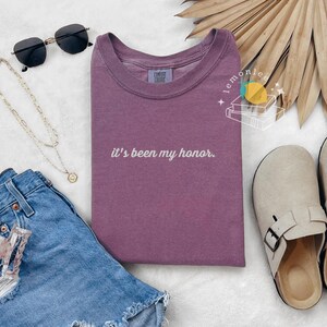 May include: A mauve t-shirt with the phrase "it's been my honor" in white script. The image also includes blue denim shorts, black sunglasses, a gold necklace, and beige slip-on clogs.