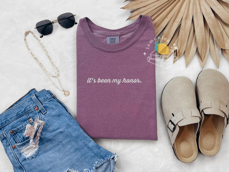 May include: A mauve t-shirt with the phrase "it's been my honor" in white script. The image also includes blue denim shorts, black sunglasses, a gold necklace, and beige slip-on clogs.