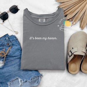May include: A casual fashion flat lay featuring a gray t-shirt with the phrase "it's been my honor" in white script, paired with denim shorts, sunglasses, a necklace, and beige sandals. A stylish, everyday outfit.