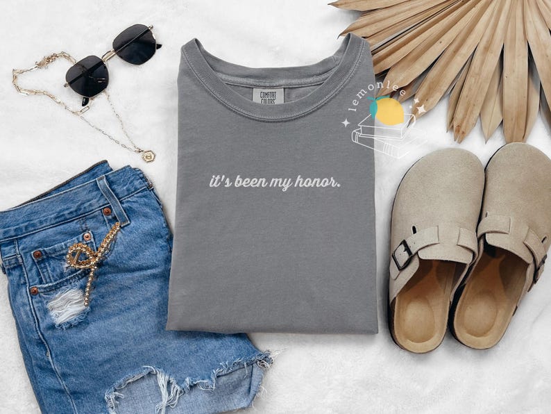 May include: A casual fashion flat lay featuring a gray t-shirt with the phrase "it's been my honor" in white script, paired with denim shorts, sunglasses, a necklace, and beige sandals. A stylish, everyday outfit.