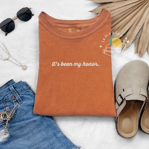 May include: An orange t-shirt with the phrase "it's been my honor" in white script. The image also includes black sunglasses, a silver necklace, denim shorts, and beige clogs.