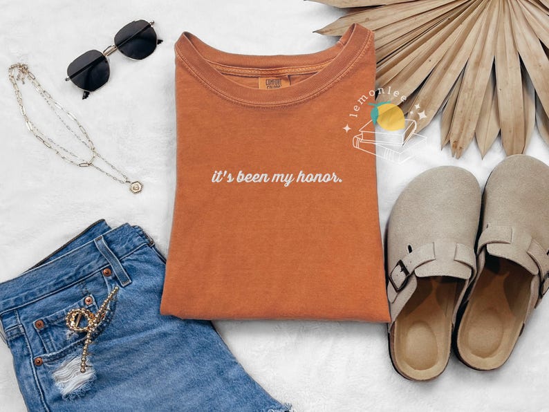 May include: An orange t-shirt with the phrase "it's been my honor" in white script. The image also includes black sunglasses, a silver necklace, denim shorts, and beige clogs.