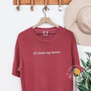 May include: A maroon-colored t-shirt with the phrase "it's been my honor" embroidered in white. The shirt is hanging on a wooden hanger. A beige hat is visible in the background.
