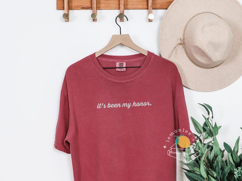 May include: A maroon-colored t-shirt with the phrase "it's been my honor" embroidered in white. The shirt is hanging on a wooden hanger. A beige hat is visible in the background.