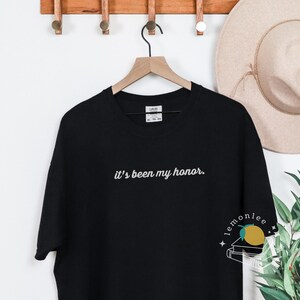 May include: Black t-shirt with the embroidered phrase "it's been my honor." The shirt is hanging on a wooden hanger. A beige hat and greenery are in the background.