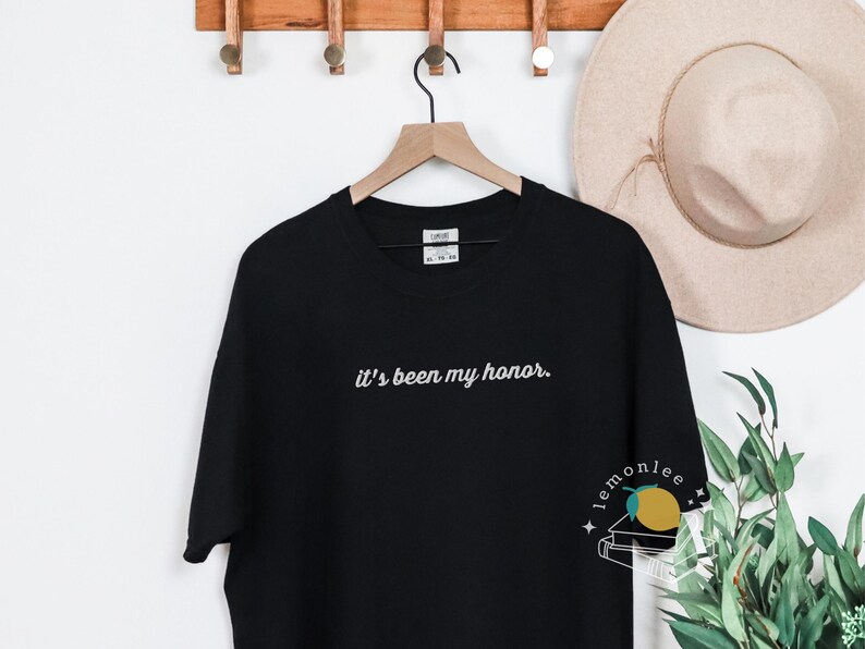 May include: Black t-shirt with the embroidered phrase "it's been my honor." The shirt is hanging on a wooden hanger. A beige hat and greenery are in the background.