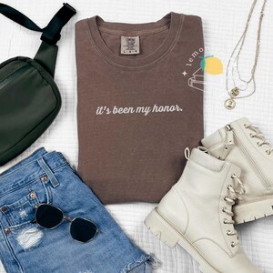 May include: A flat lay of casual apparel and accessories. Includes a brown t-shirt with the text "it's been my honor", denim shorts, white boots, a black crossbody bag, sunglasses, and layered necklaces.