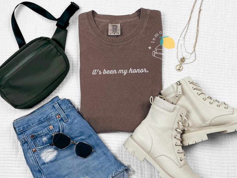 May include: A flat lay of casual apparel and accessories. Includes a brown t-shirt with the text "it's been my honor", denim shorts, white boots, a black crossbody bag, sunglasses, and layered necklaces.