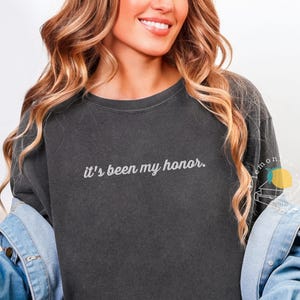 May include: Dark gray sweatshirt with the phrase "it's been my honor" in white script. The sweatshirt has a crew neck and long sleeves. The model is wearing a denim jacket over the sweatshirt.