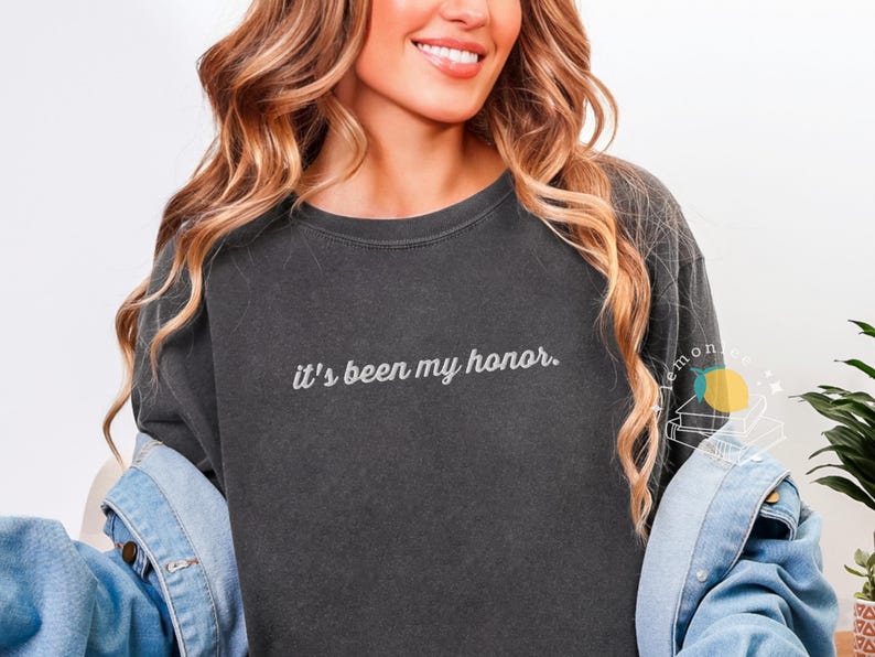 May include: Dark gray sweatshirt with the phrase "it's been my honor" in white script. The sweatshirt has a crew neck and long sleeves. The model is wearing a denim jacket over the sweatshirt.