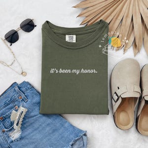 May include: An olive green t-shirt with the phrase "it's been my honor" in white script. The image also includes denim shorts, sunglasses, a necklace, and beige clogs. A dried palm leaf fan is in the background.