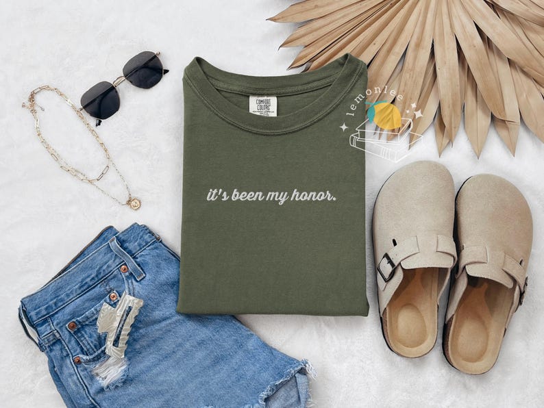 May include: An olive green t-shirt with the phrase "it's been my honor" in white script. The image also includes denim shorts, sunglasses, a necklace, and beige clogs. A dried palm leaf fan is in the background.
