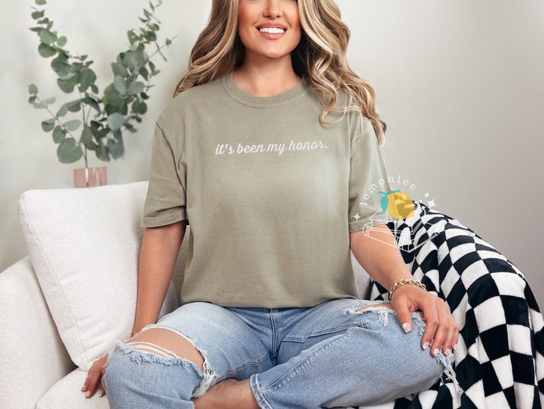 May include: Olive green t-shirt with the phrase "It's been my honor" in white script. The shirt is worn with ripped blue jeans. The model is sitting on a white couch with a black and white checkered blanket.