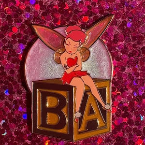 May include: A pin featuring a cartoon fairy with red hair and a red dress, sitting on a gold-trimmed block with the letters "BA". The fairy has her arms crossed and an angry expression. The pin has a glittery background.