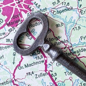 May include: A vintage metal key with a heart-shaped opening rests on a map of Berlin, Germany. The map features pink and blue lines, with place names and numbers. The key's dark, aged appearance contrasts with the map's colours.