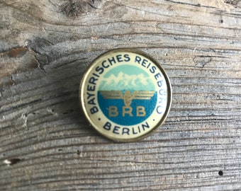 May include: A gold-colored pin with a blue and white design. The pin features a stylized eagle with the text "Bayerisches Reiseburo BRB Berlin" around the edge.