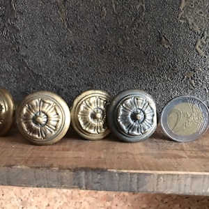 Könnte beinhalten: Fünf verzierte Messingknöpfe mit floralem Design. Die Knöpfe sind in einer Reihe auf einer Holzoberfläche angeordnet.