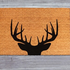 A black silhouette of a deer head with antlers is displayed on a textured orange mat against a wooden background.