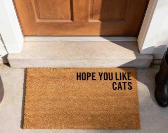 May include: A rectangular tan coir doormat with a coarse, fibrous texture. The text HOPE YOU LIKE CATS is printed in bold, black, sans-serif capital letters on the right side of the mat, positioned horizontally in two lines.