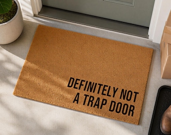 May include: A rectangular tan coir doormat with a coarse, textured surface. The mat features the black printed text "DEFINITELY NOT A TRAP DOOR" positioned in the lower right section, written in a bold, sans-serif, all-caps typeface.