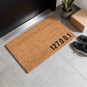 May include: A rectangular tan coir doormat featuring the black printed text 127.0.0.1 positioned in the lower right corner. The mat has a coarse, fibrous texture and is placed on a light grey tiled floor near a dark door frame.