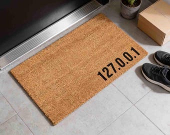May include: A rectangular tan coir doormat featuring the black printed text 127.0.0.1 positioned in the lower right corner. The mat has a coarse, fibrous texture and is placed on a light grey tiled floor near a dark door frame.