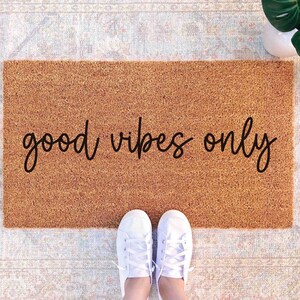 A pair of white sneakers stands on a beige area rug with the phrase "good vibes only" printed on it.