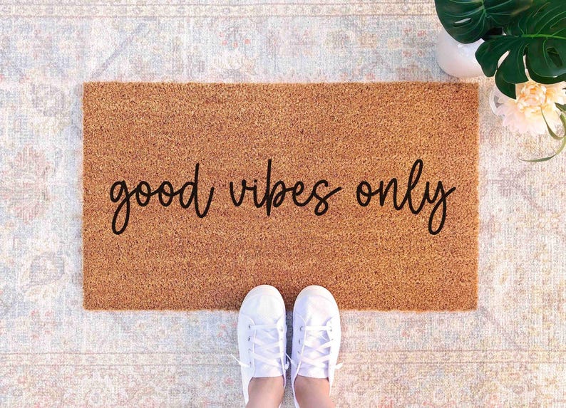 A pair of white sneakers stands on a beige area rug with the phrase "good vibes only" printed on it.