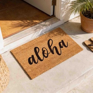 A brown doormat with the word "aloha" written on it, placed on a concrete floor next to a potted plant.