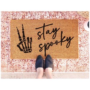 A pair of feet standing on a textured area rug with a black and white skeleton hand graphic and the text "Stay Spooky" written on it.