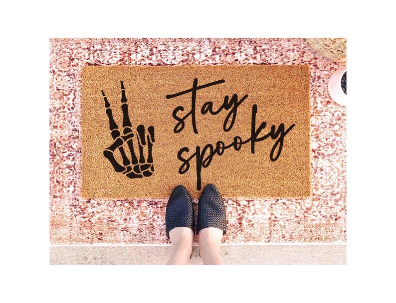 A pair of feet standing on a textured area rug with a black and white skeleton hand graphic and the text "Stay Spooky" written on it.