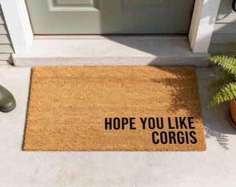 May include: A rectangular tan coir doormat with a coarse, fibrous texture. The mat features the black text "HOPE YOU LIKE CORGIS" printed in a bold, sans-serif font in the lower right corner, positioned on a concrete porch in front of a door.
