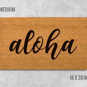 A rectangular, light brown, textured doormat with the word "aloha" written on it in a cursive font.