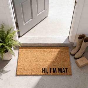 a door mat with the text "Hi, I'm MAT" on it, placed on a concrete floor next to a pair of tan boots.